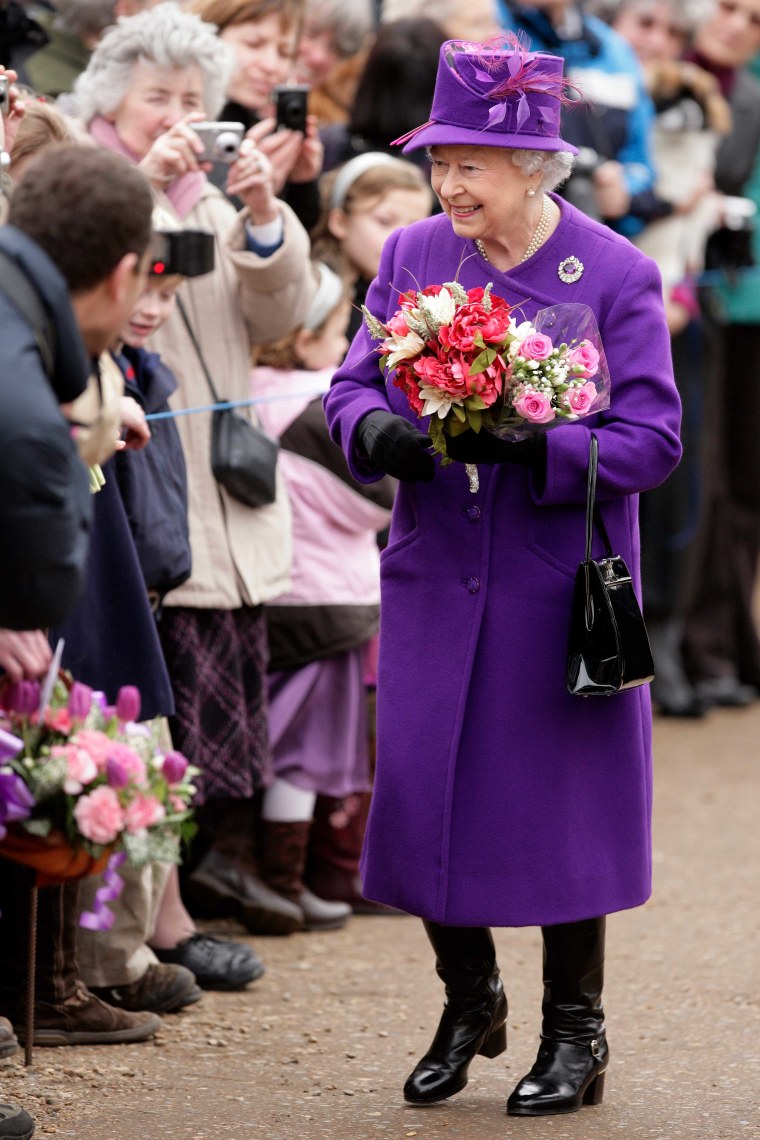Queen in a deep purple pea coat and purple top hat with a feather accent holding flowers in front of a crowd with old-fashioned digital cameras.