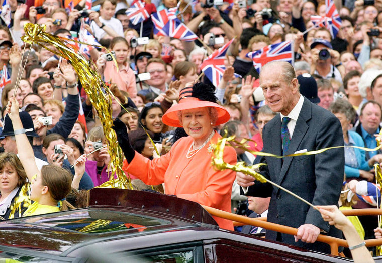 Queen Elizabeth and Prince Phillip wave from the roof of a car to people in the crowd waving flags and gold streamers.