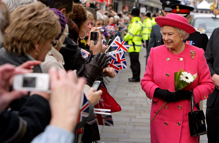 Queen Elizabeth II wears a pink jacket and matching hat as she walks down the street past crowds of people holding small British flags.