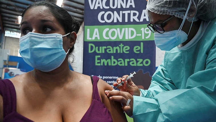 Una mujer latina sentada, con cubrebocas, es vacunada por una trabajadora médica en bata.