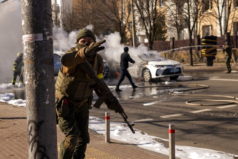 Soldados ucranianos toman posiciones en torno a una instalación militar mientras arden dos automóviles en una calle en Kiev, Ucrania, sábado 26 de febrero de 2022.