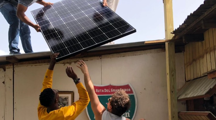Dos personas extienden sus brazos para ayudar a subir un panel solar hacia el techo de un negocio en Puerto Rico
