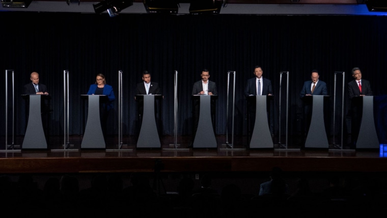 Siete políticos con traje (seis hombres y una mujer) están frente al micrófono en podios individuales separados por plexiglás durante un debate presidencial en Costa Rica.