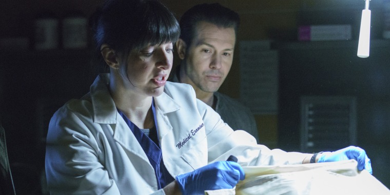 A woman with dark brown hair and bangs looks at a cadaver in a white lab coat that says medical examiner. She's wearing blue medical rubber gloves.