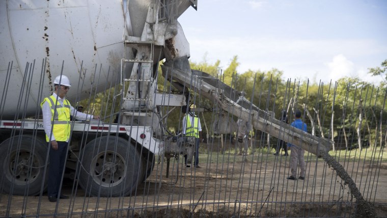 Tres personas con chaleco fosforescente están junto a un camión que carga cemento, el cual está siendo vertido alrededor de varillas que serán los cimientos de un muro fronterizo entre Haití y República Dominicana.