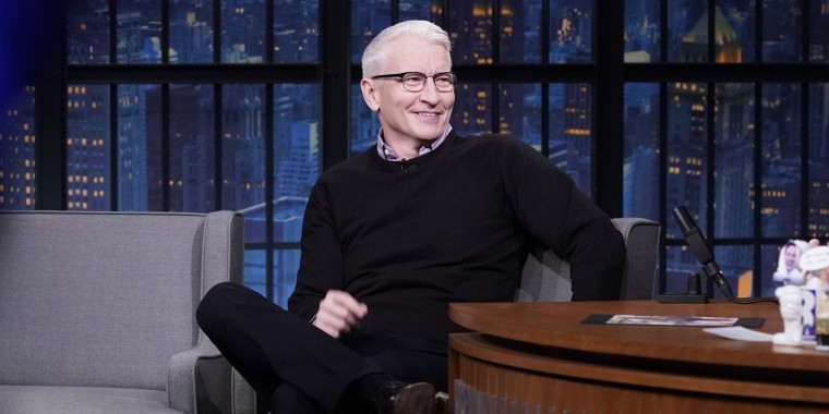 Late Night with Seth Meyers - Anderson cooper in a black sweater sits on a gray couch in front of a NYC skyline. He's smiling at Meyers off camera.