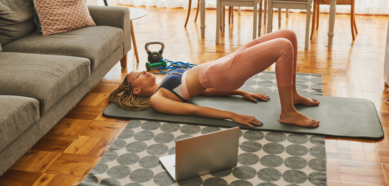 Shot of a young woman using a laptop while doing glute bridge exercises at home