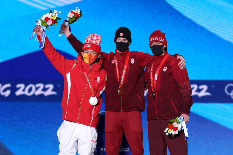 Medal Ceremony - Beijing 2022 Winter Olympics max parrot, su yiming, mark mcmorris pose on a podium in front of a blue screen holding flowers and their respective medals.