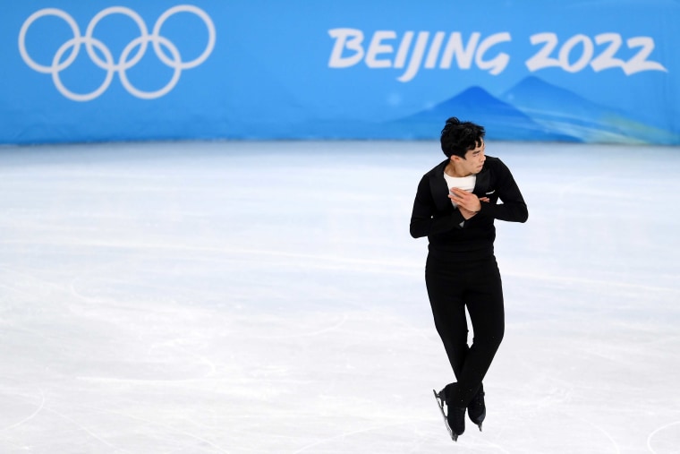 Figure Skating - Beijing 2022 Winter Olympics Day 4, Nathan Chen in all black flies through the air with his ankles crossed above the ice. A blue sign behind him says Beijing 2022.