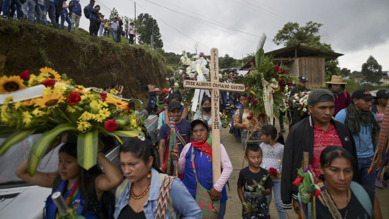 Personas caminan por un camino rural en Colombia con arreglos de flores y una cruz como parte de una procesión funeraria en honor a un líder social asesinado.