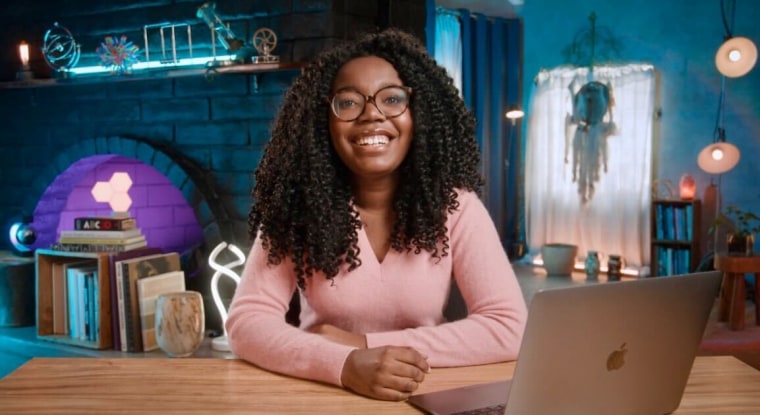 Una joven negra con anteojos sonríe en un escritorio ubicado frente a una pizarra