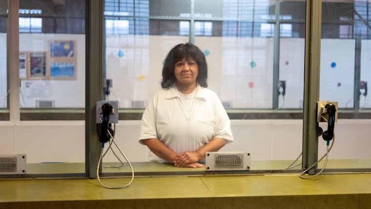 Una mujer latina parada detrás del vidrio protector de una zona de conversaciones en una prisión