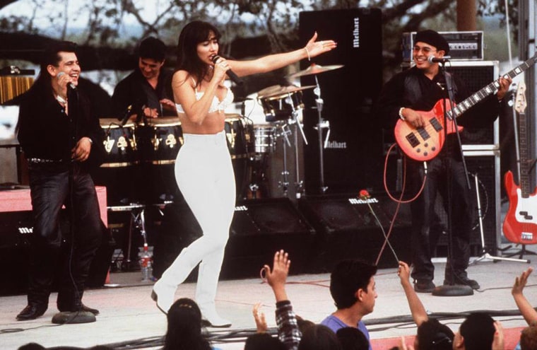 Jennifer Lopez dressed in a white bustier and tight white pants performs onstage in front of a band in all black.