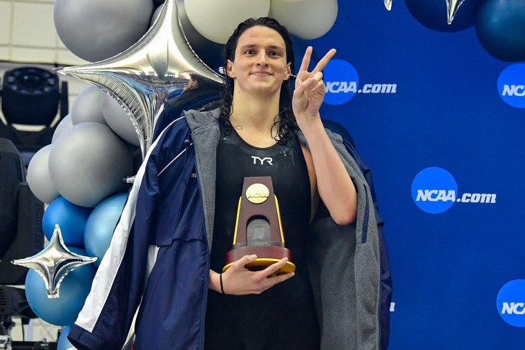 Thomas wearing a fleece-lined jacket and her swimsuit holds a wooden trophy while holding up a peace sign in front of blue and silver balloons and a step-and-pose wall with the NCAA logo.