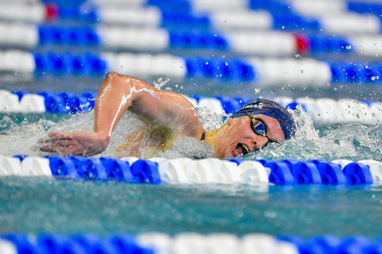Lia Thomas in a blue swim cap and mirrored goggles takes a breath as she swims freestyle in a lane framed by blue and white lane lines.