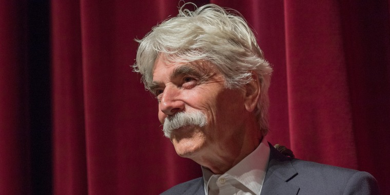 Sam Elliott in a gray suit with his signature white hair and mustache stands in front of a red curtain.