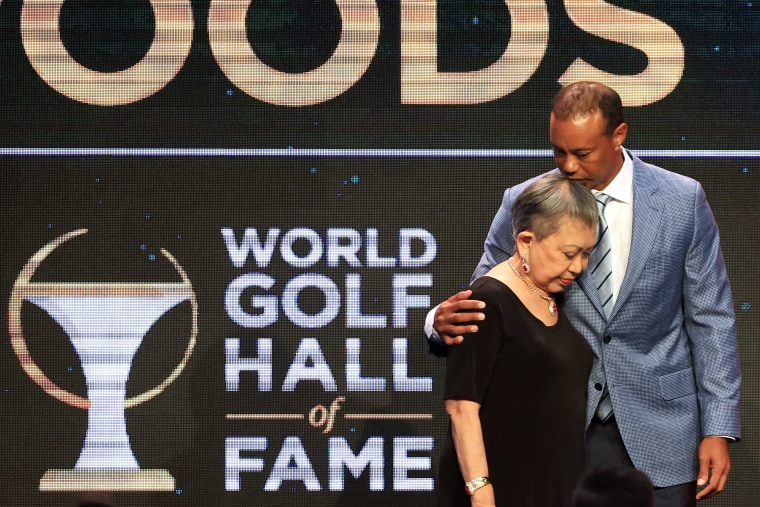 Tiger woods kisses the top of his mom's head as they stand in front of a black and gold screen.
