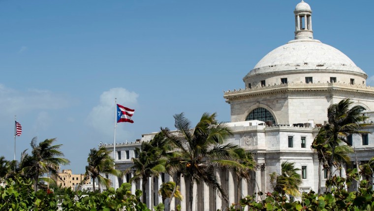 Palmeras y una bandera puertorriqueña se mueven en el viento frente a un edificio domado blanco, el Capitolio boricua