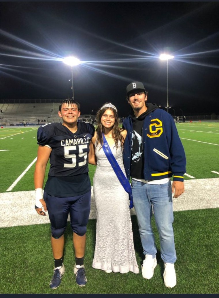 Un adolescente en uniforme de futbol americano, una joven con corona de reina de fiesta Homecoming, y un joven con gorra y chaqueta de UCLA sonríen a la cámara