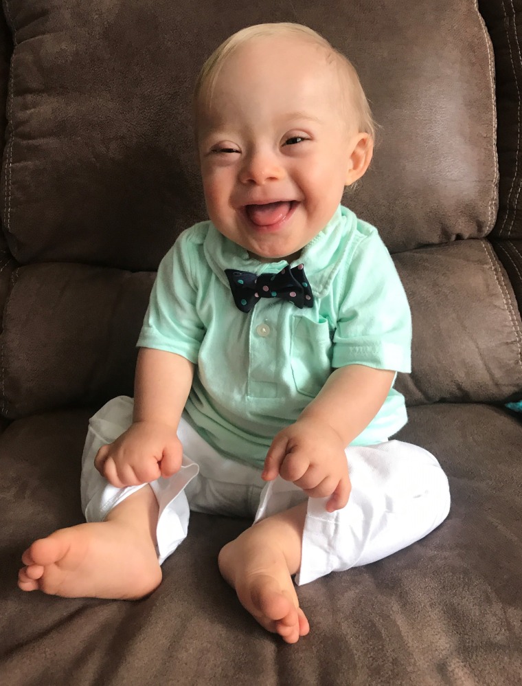 A barefoot blonde baby with Down syndrome in a mint green shirt and bow tie sticks his tongue out at the camera while grabbing the legs of his white pants.