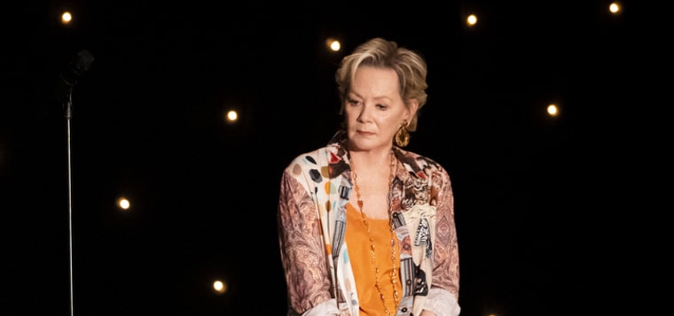 Jean SMart in a multicolored shirt and orange tank sits on a dark stage, looking off to the right with a serious expression