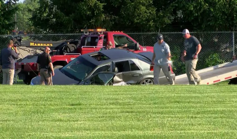 A silver sedan with clear damage to the driver side door is loaded onto a tow truck. Four men stand nearby, one in a Sheriff's office uniform.