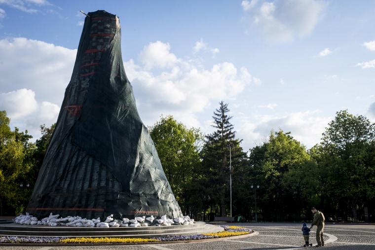 A child and a man in a military uniform pass a monument to Ukrainian Poet Taras Shevchenko that has been surrounded by protective sandbags on May 18, 2022, in Kharkiv.