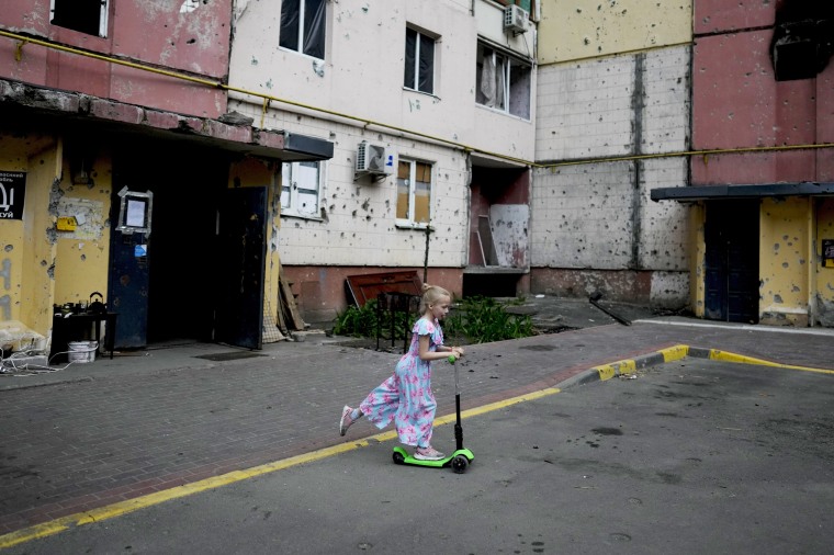 A girl rides a scooter near destroyed buildings during attacks in Irpin