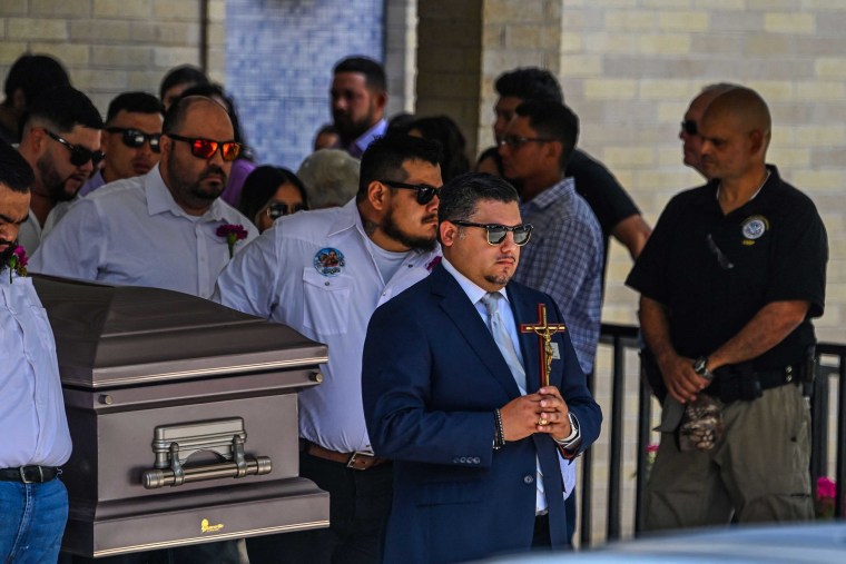 Pallbearers in white shirts carry a silver casket out of a church. They follow a man in a navy suit and Ray-ban sunglasses carrying a small wooden and metal cross with a Jesus figurine on it.