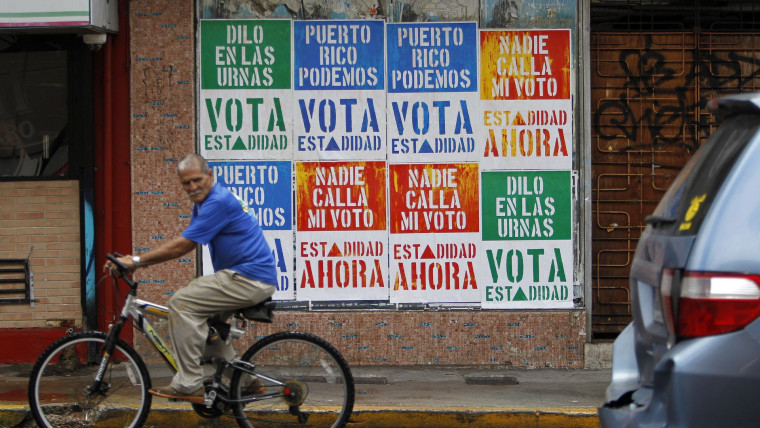 Un hombre en bicicleta pasa enfrente de un muro con carteles que dicen "Vota estadidad" antes de un referendo de 2017