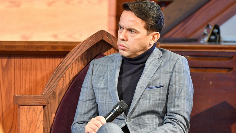 El reverendo Samuel Rodríguez Jr., hombre latino con traje sastre gris, tiene micrófono en mano mientras está sentado dentro de una iglesia