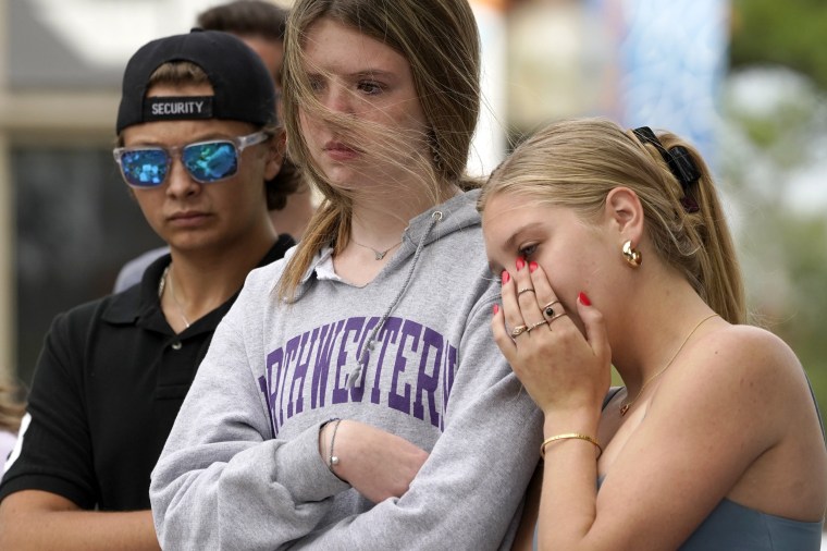 Image:  Helena Kavanaugh, right, stands with friends Addison Schwan, center, and Charlie Shookman after Kavanaugh placed flowers at a memorial for the seven people who lost their lives in the Fourth of July mass shooting in Highland Park. on July 6, 2022.