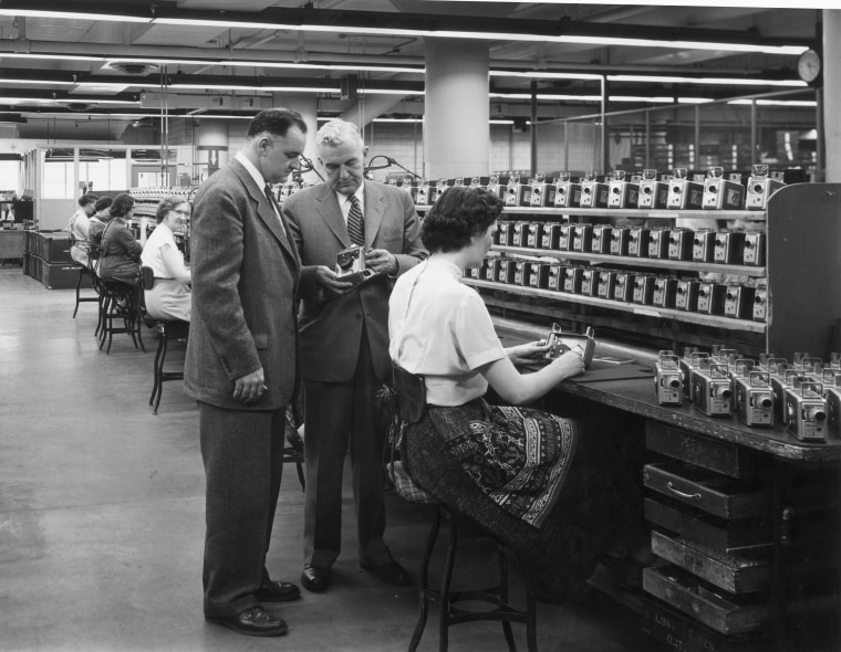 A line of female employees work on an assembly line for the Eastman Kodak Brownie camera, as two male supervisors inspect one of the cameras, circa 1945. The finished cameras line shelves above the bench.