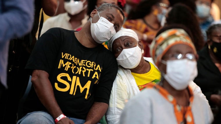 Un hombre con mascarilla facial reposa su cabeza sobre una mujer. El hombre trae una playera que dice "Vidas negras importam," el lema de Black Lives Matter en portugués.