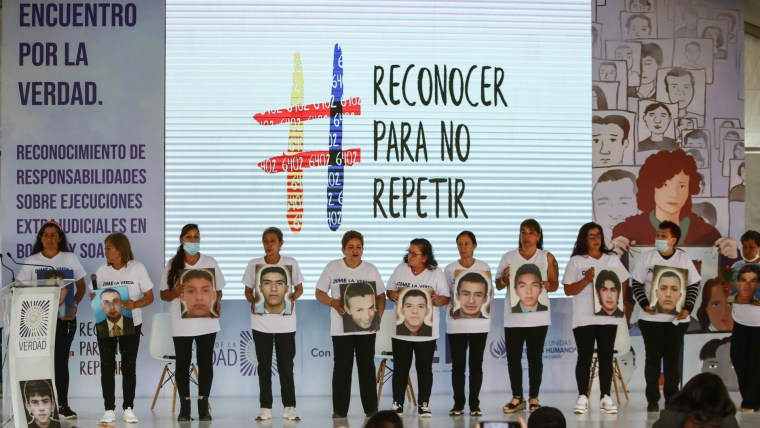 Diez mujeres vestidas de blanco posan con el retrato de sus hijos frente a una pantalla que dice "Reconocer para no repetir" en Colombia