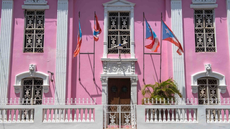 Cuatro banderas puertorriqueñas ondean en la fachada de un edificio tipo colonial de color rosa brillante