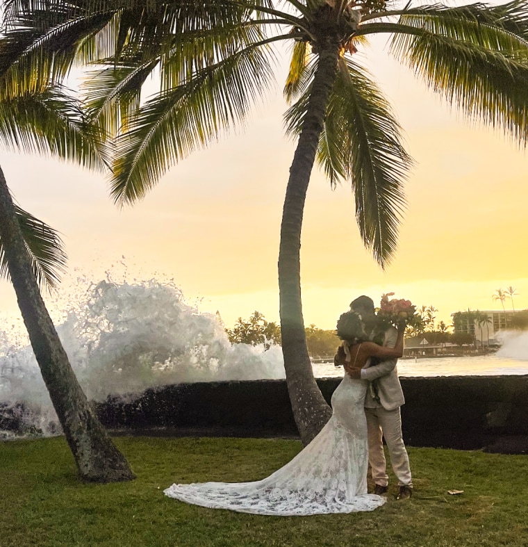A bride and groom kiss in front of a sunset and giant wave crashing over a sea wall