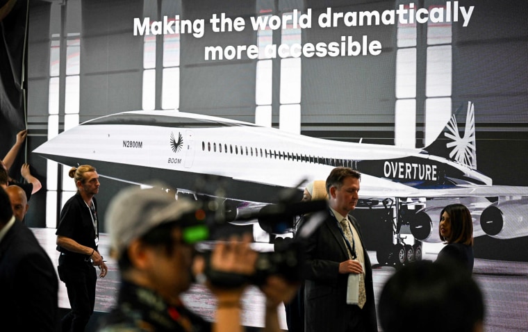 Attendees walk past a poster of a Boom Supersonic concept Overture aircraft at the Farnborough Airshow, in Farnborough, England, on July 18, 2022.