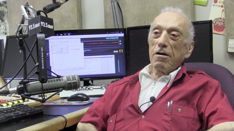 Un hombre de camiseta roja está sentado frente a un micrófono de radio y controles de transmisión