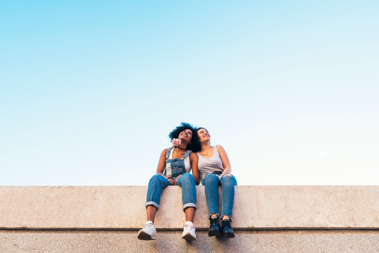 Low Angle View Of Female Friends Taking Selfie While Sitting On Retaining Wall