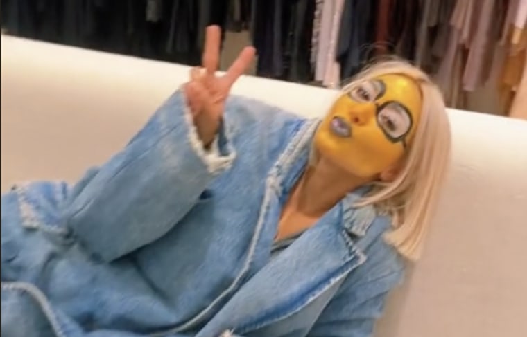 Kardashian poses in a jean blazer on a white couch with yellow face paint and black painted on glasses. her hair is styled in a blond bob.
