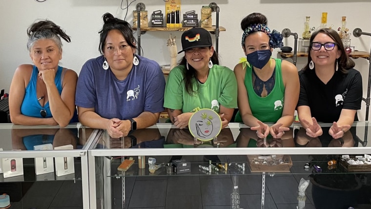 Cinco mujeres latinas sonríen hacia la cámara detrás de un aparador de vidrio en el dispensario de cannabis La Tiendita de Motita