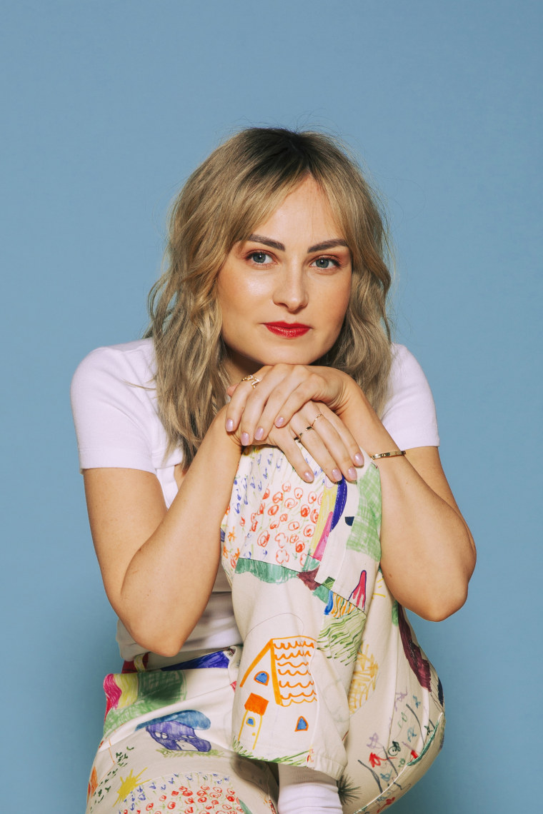 Molly Burke, a white woman, wearing pastel rainbow pants, a pink shirt, stares at the camera.