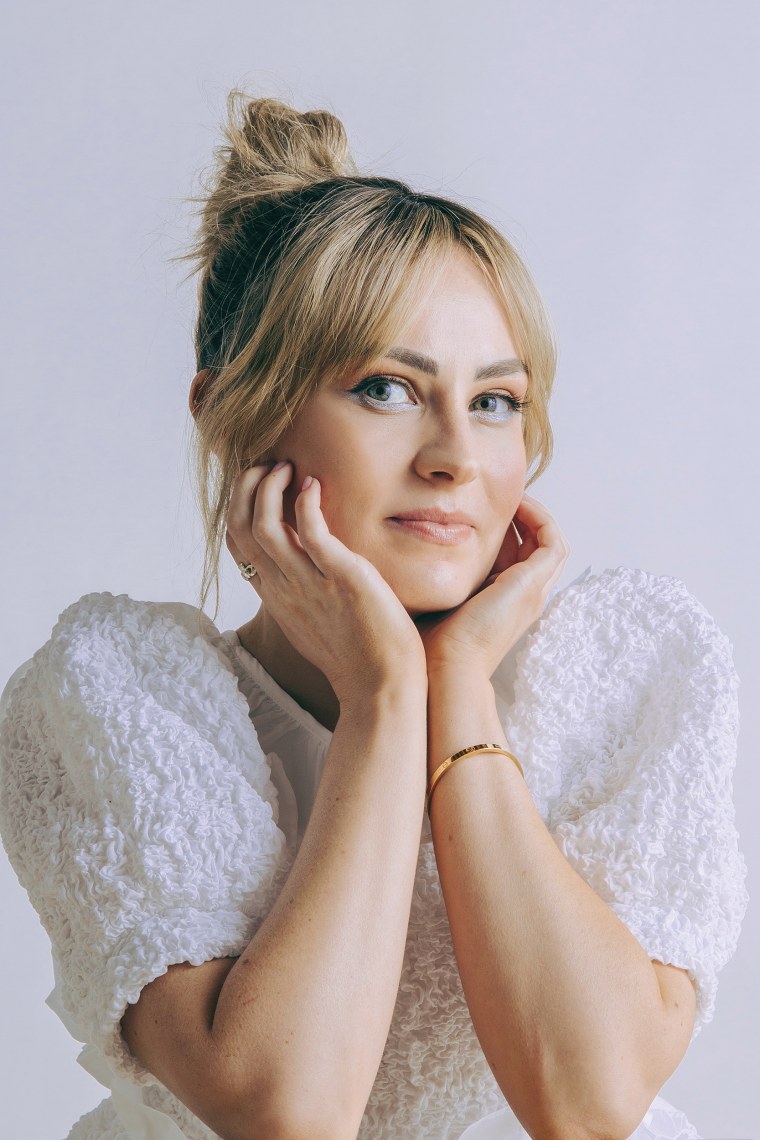 Molly Burke, a white woman, gazes at the camera. She's wearing a white dress with puffy sleeves with her blonde hair pulled back in a knot.