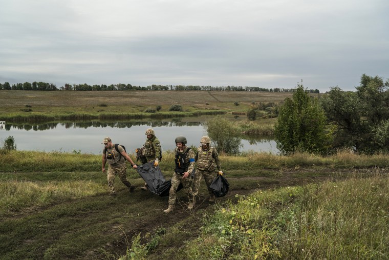 Image: Ukrainian servicemen carry a bag containing the body of a Ukrainian soldier, center, as one of them, right, carries the remains of a body of a Russian soldier in a retaken area near the border with Russia in Kharkiv region, Ukraine, on Sept. 17, 2022.