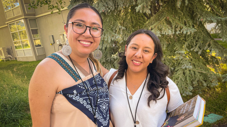 Dos mujeres latinas posan frente a un pino. A la izquierda está Liz Velasco, aspirante al senado estatal de Colorado, y del lado derecho con camisa blanca está Jasmin Ramirez, integrante de un puesto electo en una junta escolar