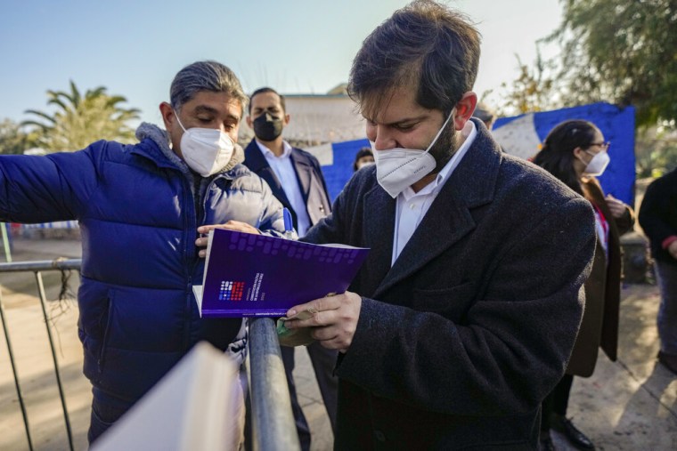 El presidente de Chile, Gabriel Boric, como siempre sin corbata, hojea un cuaderno morado que tiene una copia del borrador de una nueva constitución mientras un simpatizante lo saluda, el 21 de julio de 2022