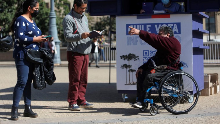 Una mujer vestida de azul está parada junto a un hombre de chaqueta gris hojeando una copia del borrador para una nueva constitución chilena mientras hablan con un hombre en silla de ruedas sobre el texto