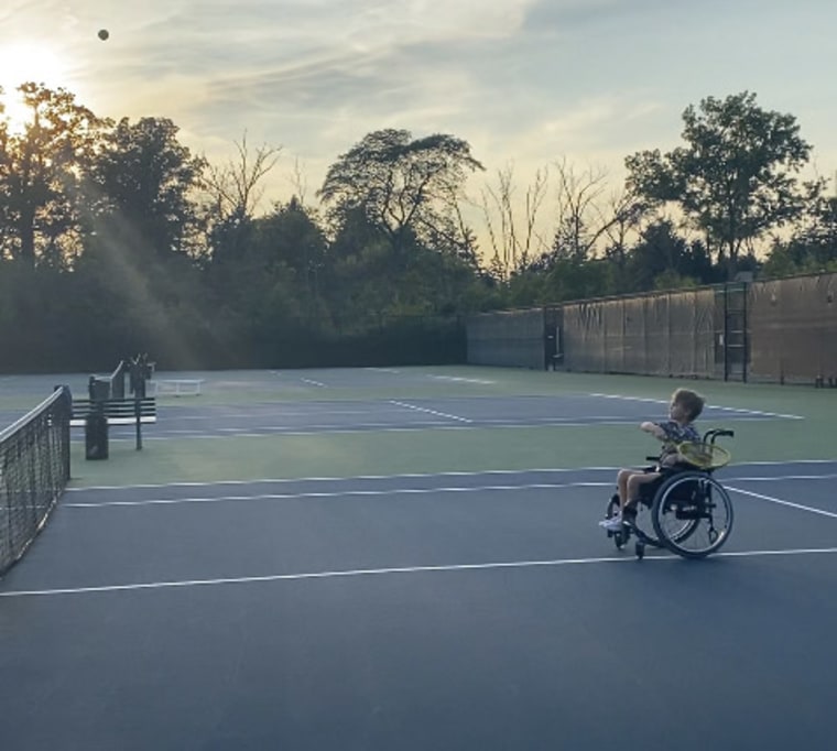 A little boy in a wheelchair swings a yellow racket on a tennis court as the sun sets.
