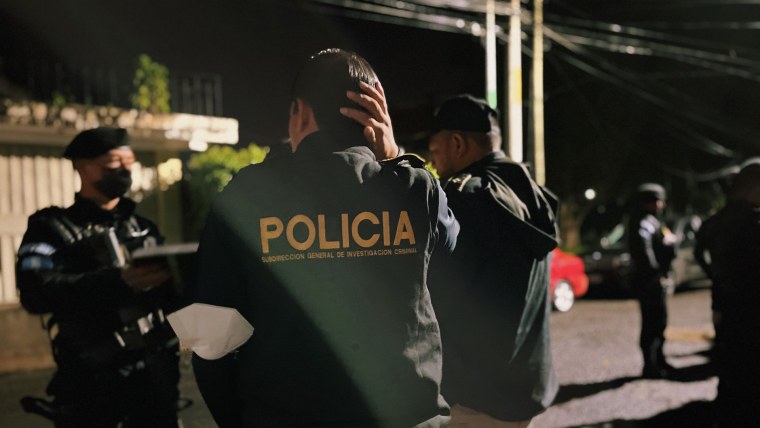 Tres agentes policiales guatemaltecos con uniforme y mascarilla hablan en una calle debajo de una luminaria pública antes de un allanamiento